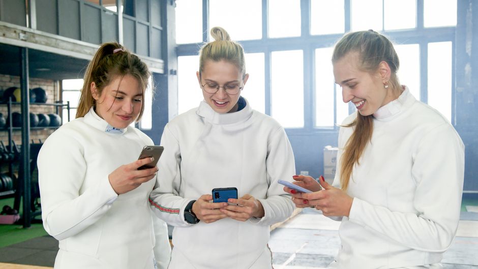 Three female fencers in a gym smiling and using smartphones after training.
